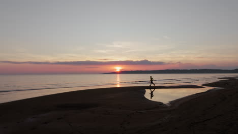 aerial: silueta de un hombre corriendo a lo largo de la orilla de la playa durante la puesta de sol. playa de arena de kavouri, katakolo, provincia de elis