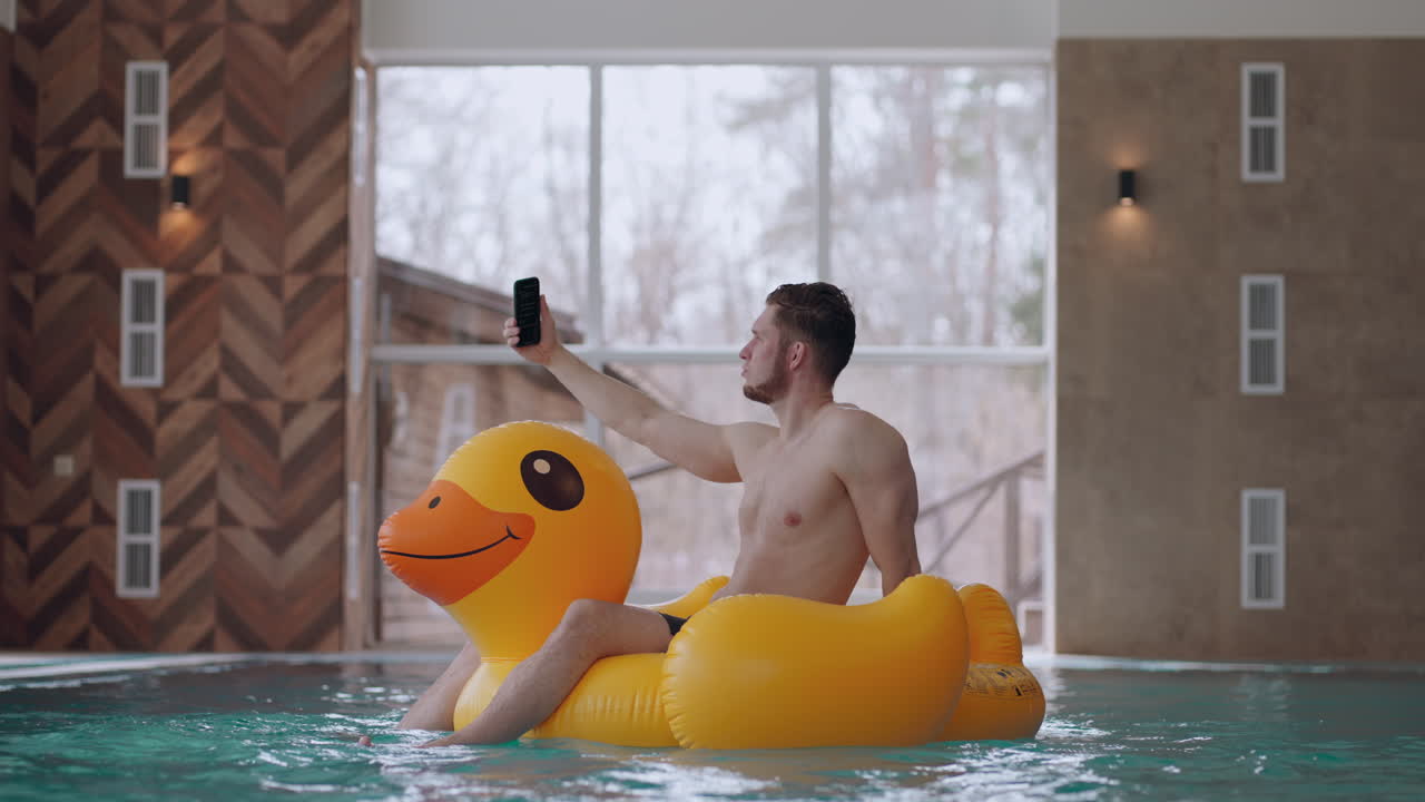 adult man is resting in swimming pool in water park floating on funny inflatable duck and taking selfie by smartphone