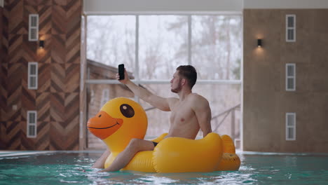 Erwachsener-Mann-Ruht-Sich-Im-Schwimmbad-Im-Wasserpark-Aus,-Schwimmt-Auf-Einer-Lustigen-Aufblasbaren-Ente-Und-Macht-Ein-Selfie-Mit-Dem-Smartphone