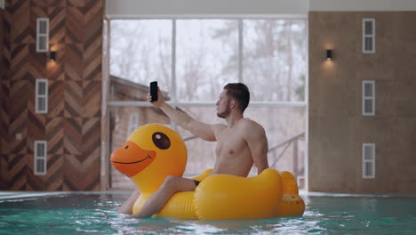 un hombre adulto está descansando en la piscina de un parque acuático flotando en un pato inflable divertido y tomando una selfie con un teléfono inteligente