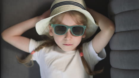 sad little girl in sunglasses with green rim lies on sofa