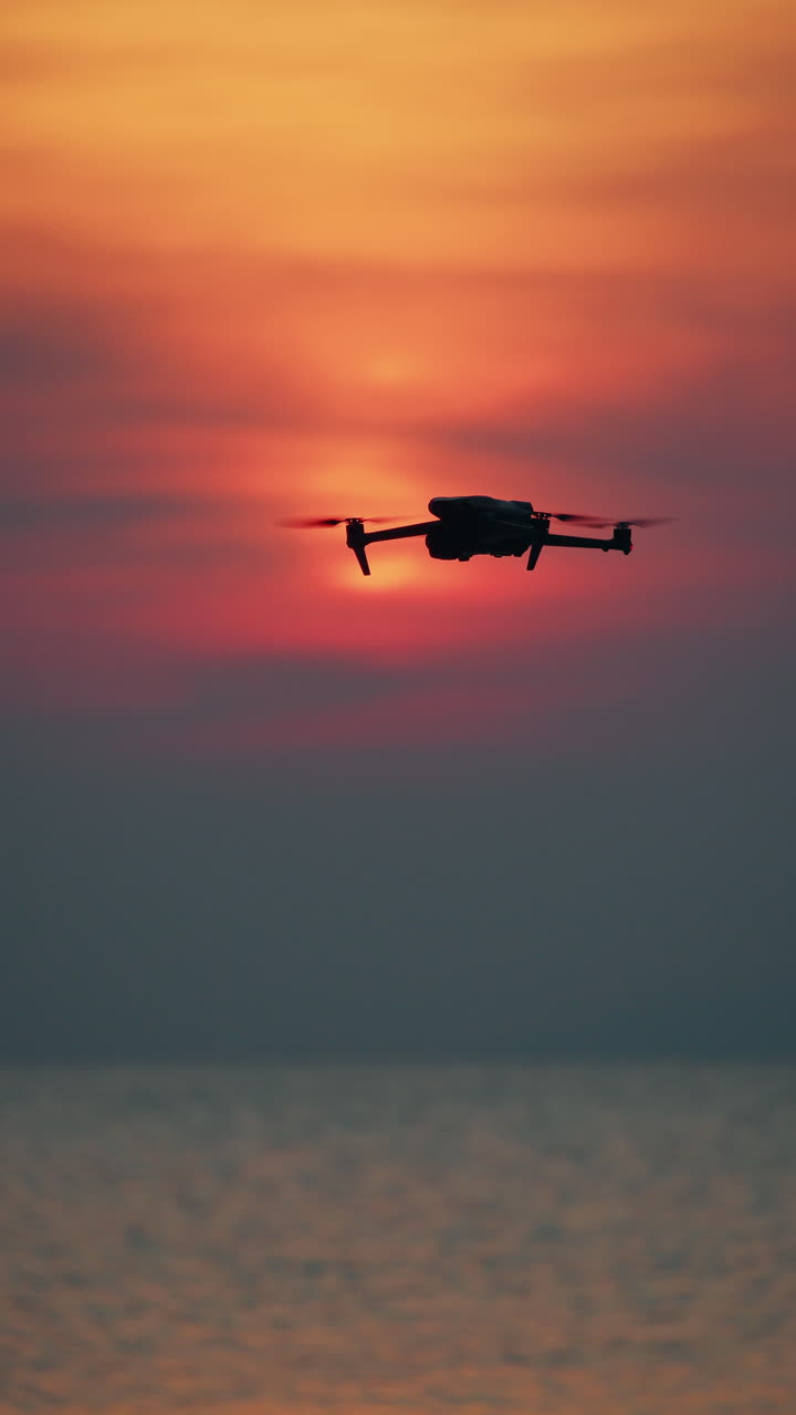 Dronesilhuet svæver over havet ved solnedgang med blød rødorange himmel.