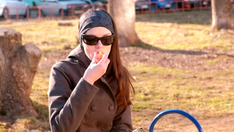 young brunette woman in sunglasses eats tangerine sitting on the bench in park.