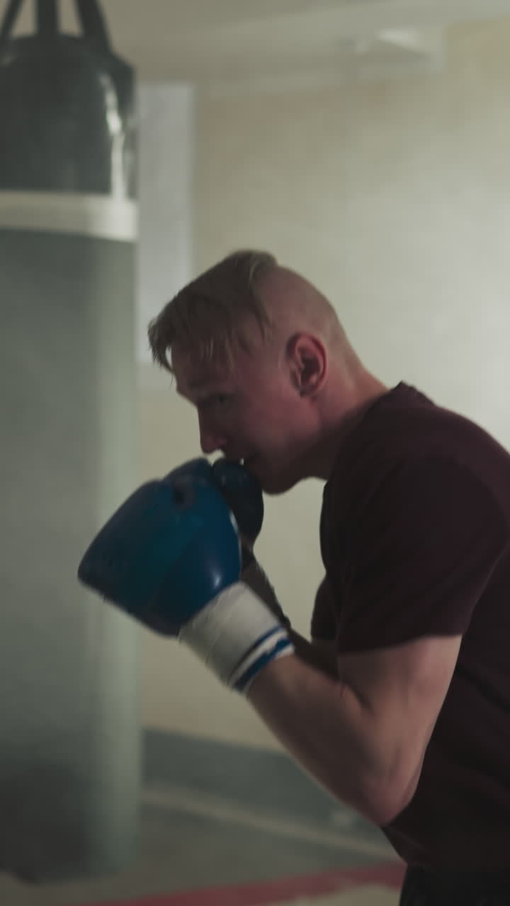 Boxer punches heavy bag exercising in sports center. Emotional athlete in gloves beats hanging item by fists in gym slow motion. Relax technique