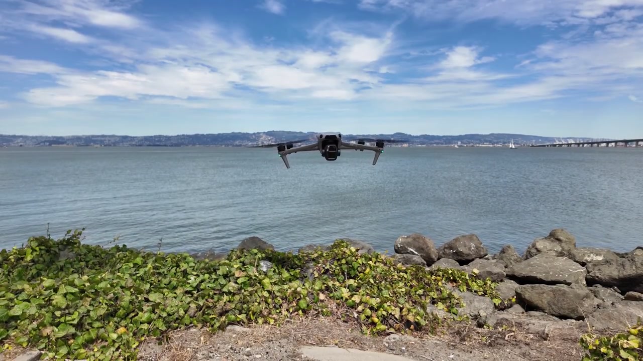 Moderno drone per consumatori che sorvola la costa rocciosa prima di allontanarsi sulla Baia di San Francisco, con il Bay Bridge e le colline dell'East Bay visibili sullo sfondo sotto un cielo blu.
