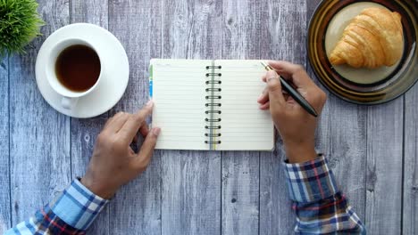 person, die in einem notizbuch mit kaffee und croissant schreibt