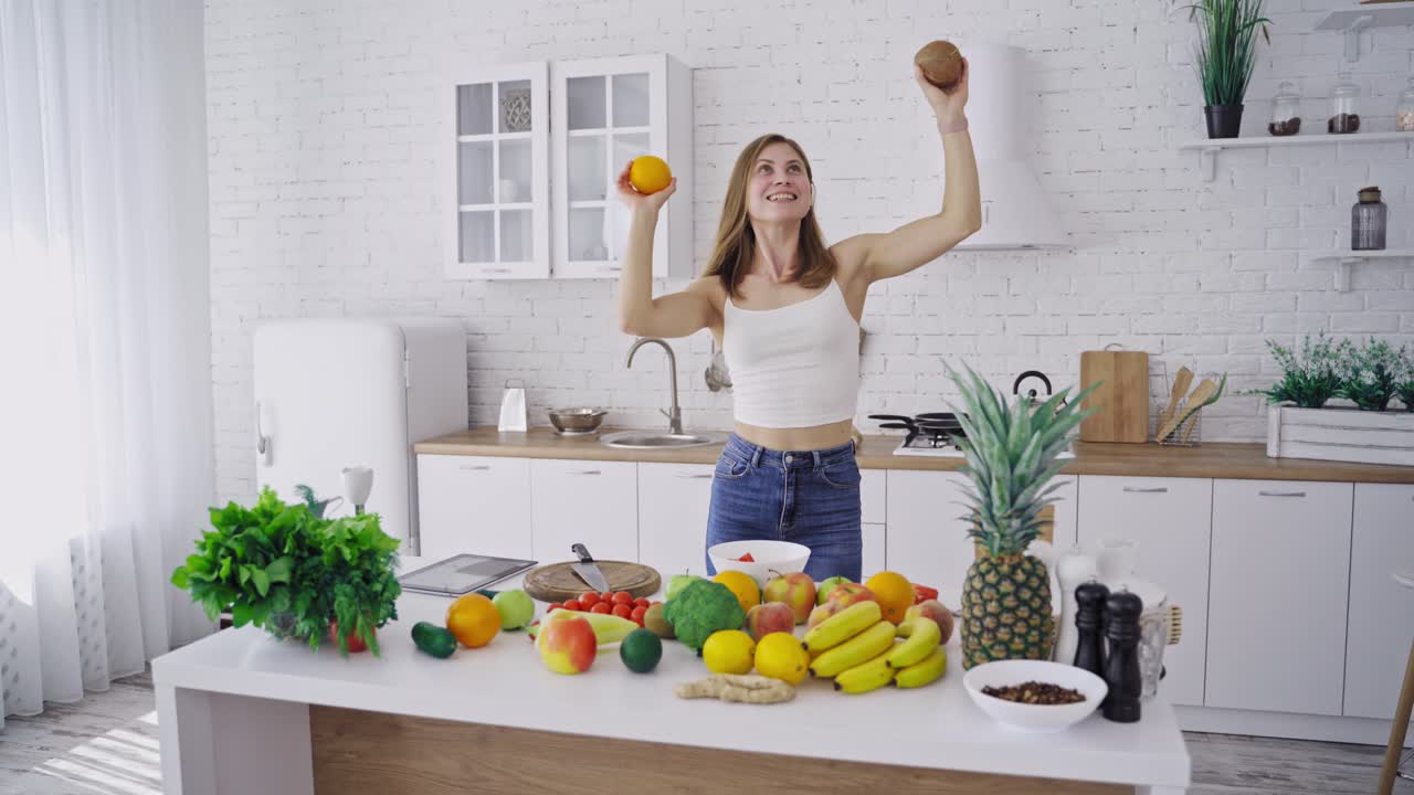 Beautiful slim woman is dancing with fresh fruits in the modern kitchen. Happy woman holding coconut and orange having fun at home. Organic fruit and vegetables. Dieting concept.