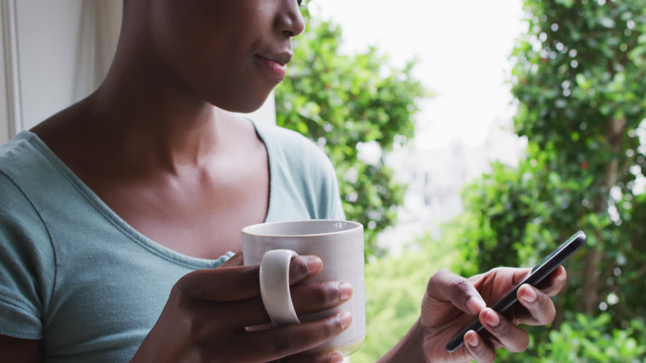 mujer afroamericana sosteniendo una taza de café usando un teléfono inteligente en casa
