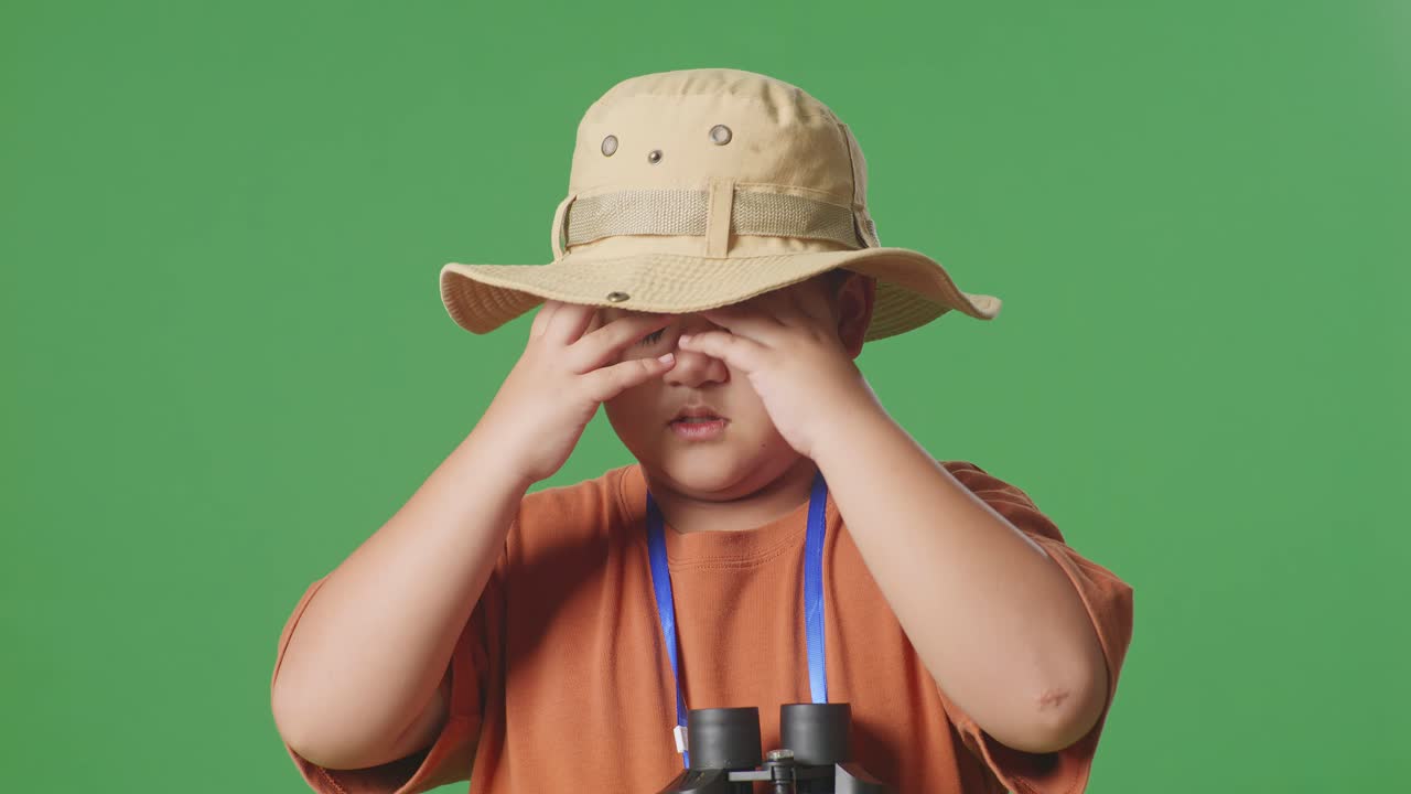 모자를 입은 아시아 관광 소년은 안경을 통해 보고 두통을 고 있습니다. 소년 연구자는 녹색 화면 배경에서 무언가를 조사합니다. 여행 관광 모험 개념, 근접