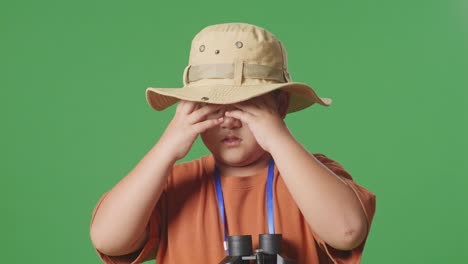 niño turista asiático con un sombrero que tiene dolor de cabeza después de mirar a través de los binoculares. niño investigador examina algo en el fondo de la pantalla verde, concepto de aventura de turismo de viaje, de cerca
