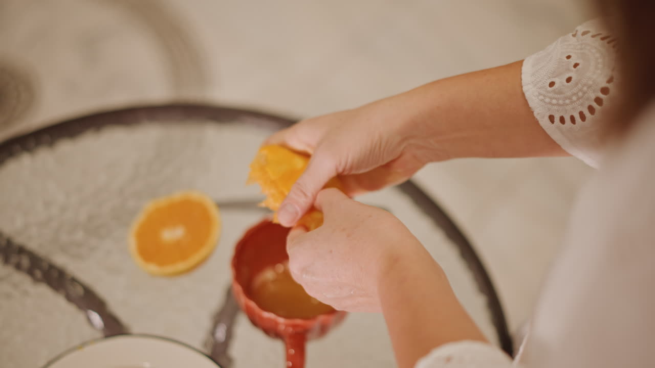 Manos desde arriba exprimiendo naranja sobre una taza. Mesa de cristal con una rodaja de naranja y una taza de cerámica, reflejo suave de la luz solar, vertido delicado en una bebida caliente, ritual de desayuno casero, estilo de vida acogedor.