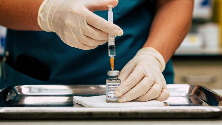 Steadying vial, gloved nurse in teal scrubs at counter lowering syringe drawing amber dose for shot