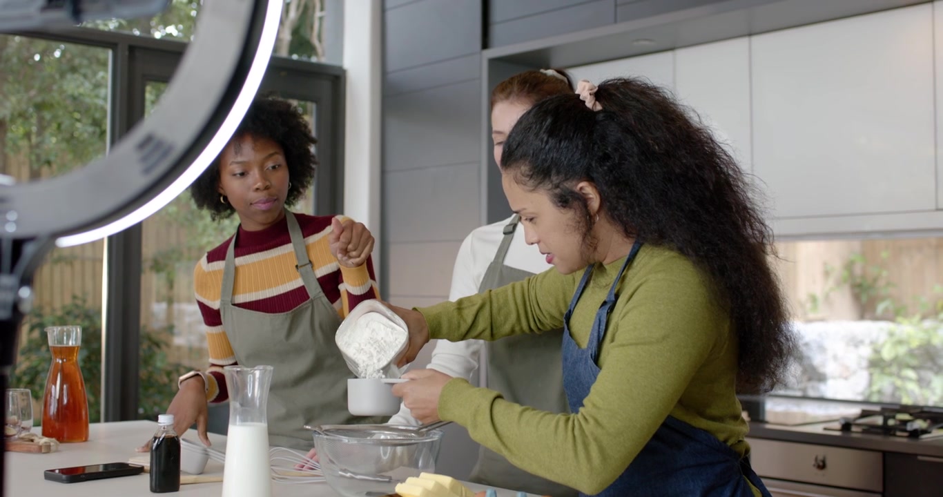 Mulheres diversas de avental observando a mulher à direita segurando uma colher medindo farinha em uma tigela, filmando receita.