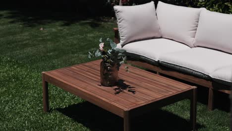 wooden outdoor table with a vase of eucalyptus leaves beside a soft cushioned bench in sunlight