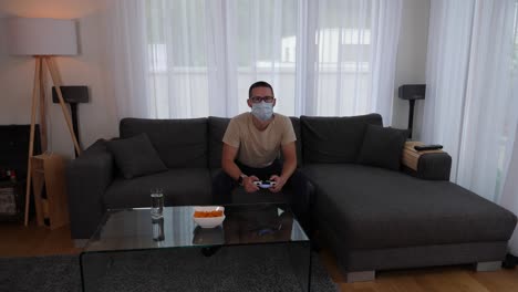 young man with face mask sitting on sofa playing video games