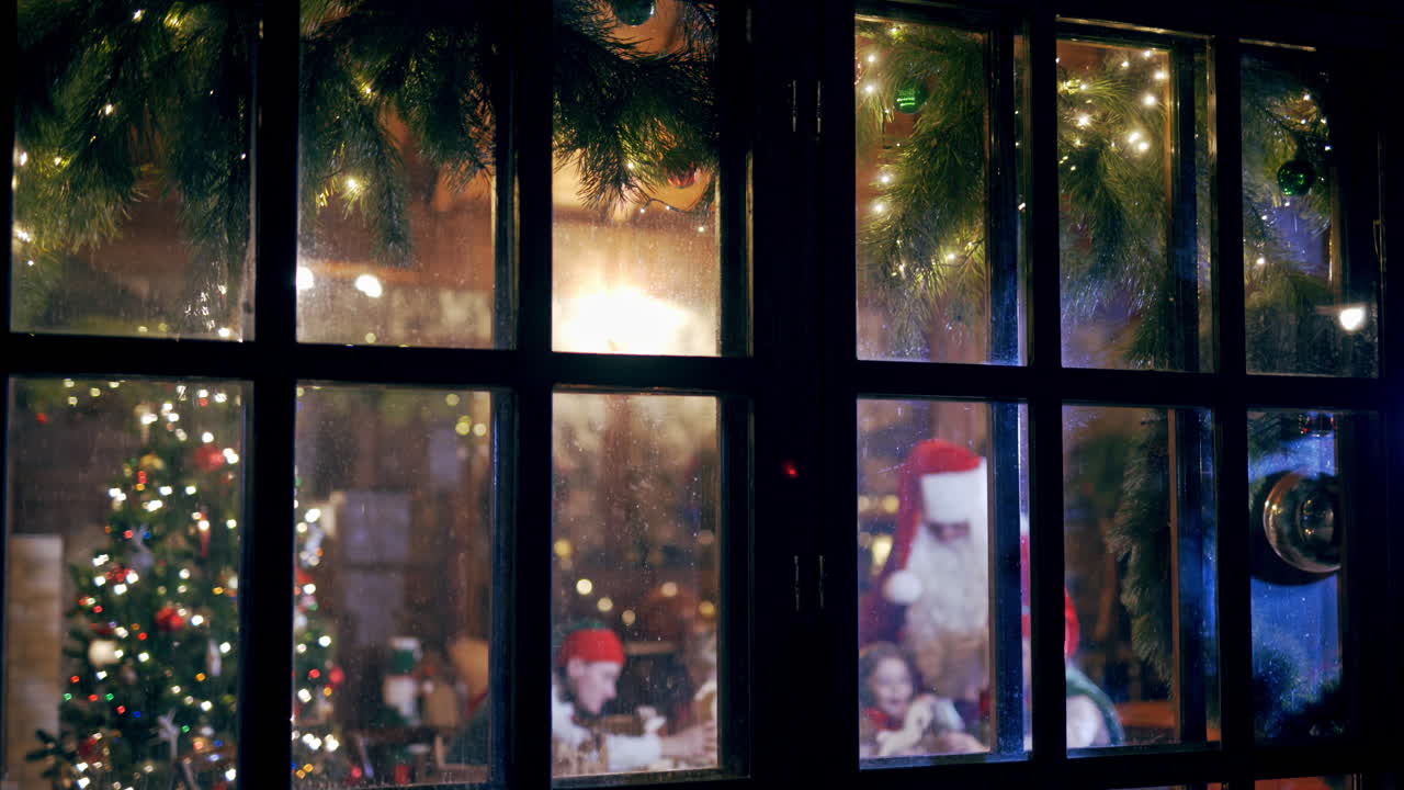 Christmas decorations, happy children and Santa Claus inside the house. Santa is talking to children and elves in the house. Christmas atmosphere through the large window in the evening.