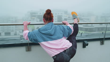 Sporty-fit-woman-in-blue-pink-sportswear-doing-squats-yoga-stretching-exercising-on-roof-of-house