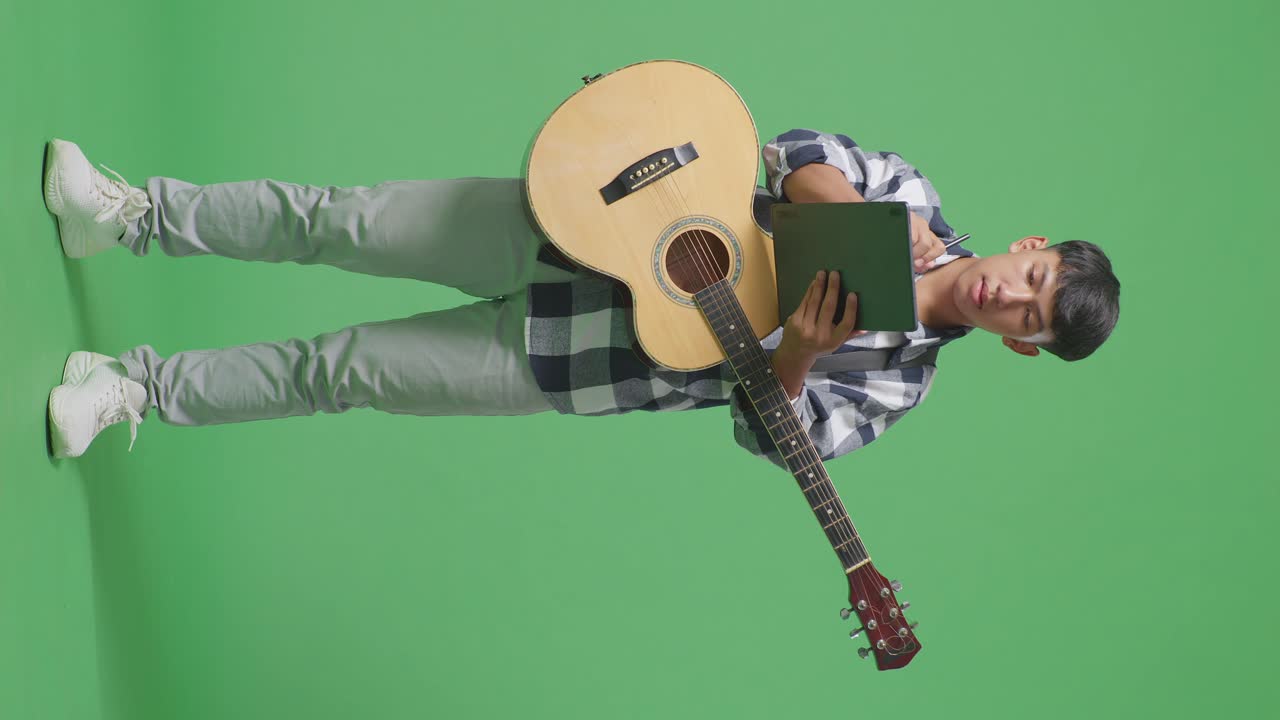 Adolescente asiatico a figura intera con chitarra che prende appunti su un tablet mentre è in piedi su sfondo green screen.