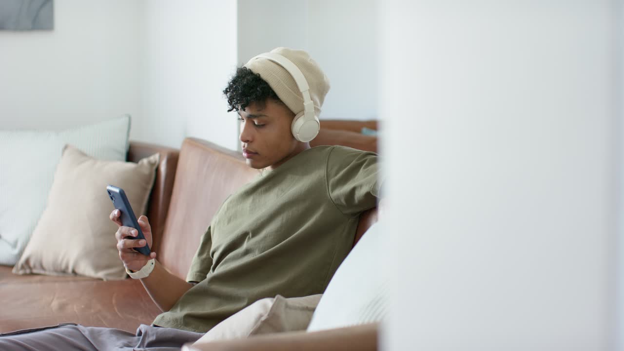 African American man early 20s checking smartphone, placing white headphones, scrolling on sofa