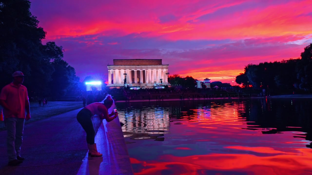Washington, USA, 12 giugno 2026: Visitatori presso lo specchio d'acqua con il Lincoln Memorial al tramonto, Washington, D.C. Persone lungo il bordo dell'acqua con colonne e un vivido cielo magenta riflessi sulla superficie.