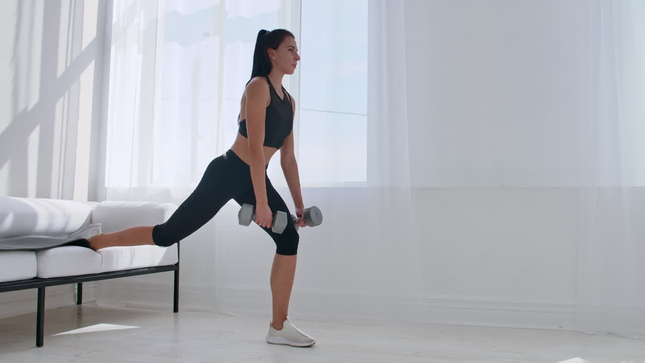 Una bruna in abbigliamento sportivo nero in un appartamento bianco esegue uno split squat con manubri tra le mani, appoggiando il piede sul divano.