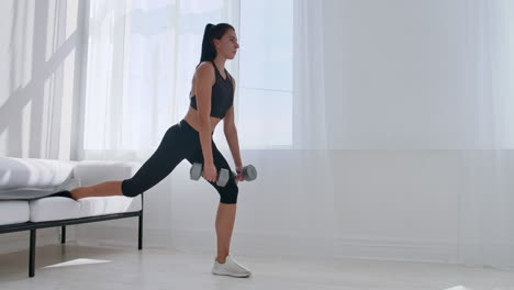 brunette in black sportswear in a white apartment makes a split squat with dumbbells in her hands leaning on the sofa with her foot.