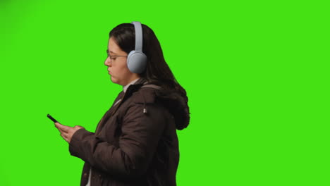 young woman wearing wireless headphones streaming music from mobile phone walking across frame against studio green screen