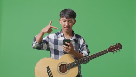 초록색 스크린 배경에서 손에 스마트폰을 가리키는 기타를 들고 있는 아시아 십대