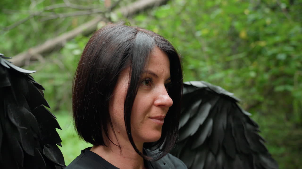 Black fairy with dark wings standing quietly in green forest, head bowed in calm reflection, soft light touching black feathers and dark hair, creating serene mystical mood