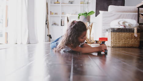 Young-Hispanic-girl-lying-on-the-floor-in-the-sitting-room-playing-with-toy-digger-truck,-front-view