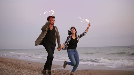 young happy multi ethnic couple holding burning sparkling candles and running by the sea during sunset. slow motion shot