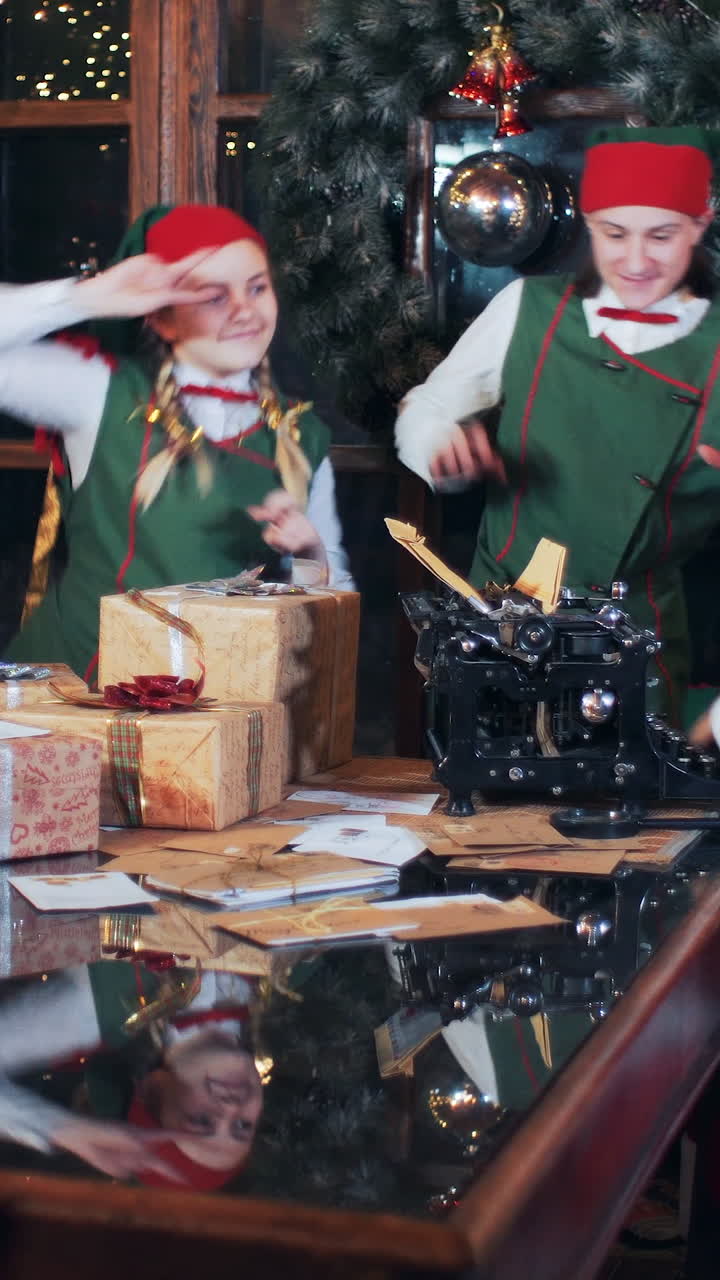 Santa Claus is sitting at a table with candles and an old typewriter in a room with New Year's decorations. Elves in green costumes are dancing. Vertical video