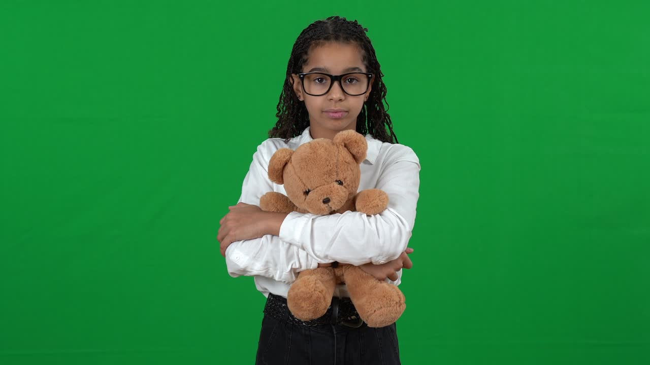 Portrait of cute unhappy African American teen girl hugging teddy bear standing on green screen. Sad nerd teenager in eyeglasses posing at chromakey background looking at camera.