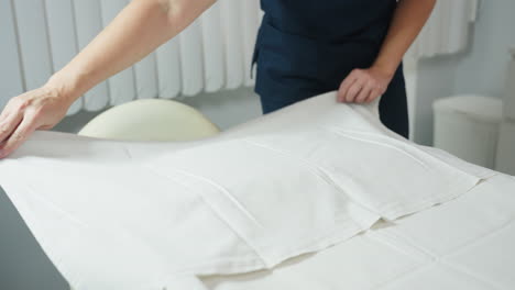 facialist in navy uniform smooths crisp white cloth over cream massage chair from low perspective in bright white treatment room, preparing hygienic, comfortable surface for client facial