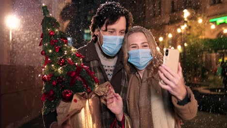 caucasian happy couple wearing facial masks holding christmas tree and waving while making a video calle with smartphone on the street in christmas