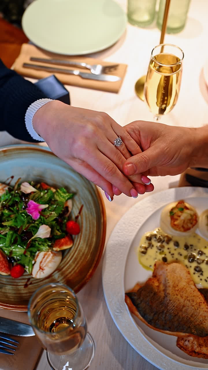 Una coppia si gode una cena e festeggia il fidanzamento con cibo e gioia. Due mani si tengono sopra una tavola imbandita con piatti e bevande.