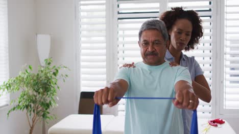 Fisioterapeuta-Femenina-De-Raza-Mixta-Ayudando-A-Un-Hombre-Mayor-A-Hacer-Ejercicio-Usando-Una-Banda-De-Ejercicio