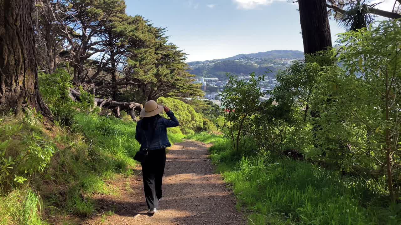 Una donna con una giacca di jeans e un cappello da sole cammina per esplorare un sentiero nel bosco e scopre una vista della città di Wellington, Nuova Zelanda