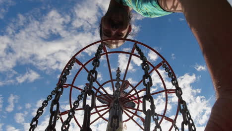 a unique perspective from inside the disc golf basket looking up at a man reaching in, set against a bright blue sky with clouds