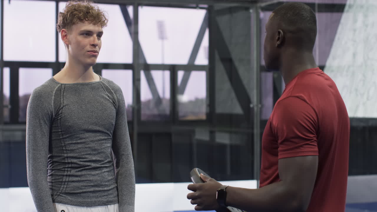 Two diverse men enjoying padel tennis indoors, holding rackets and smiling, at indoor court