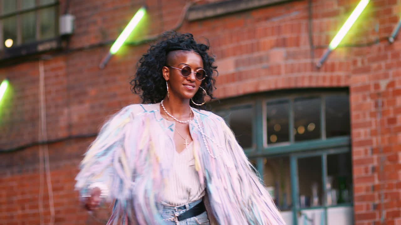 Fashionable young black woman wearing multicoloured fringed jacket, sunglasses and jeans turning around laughing, low angle