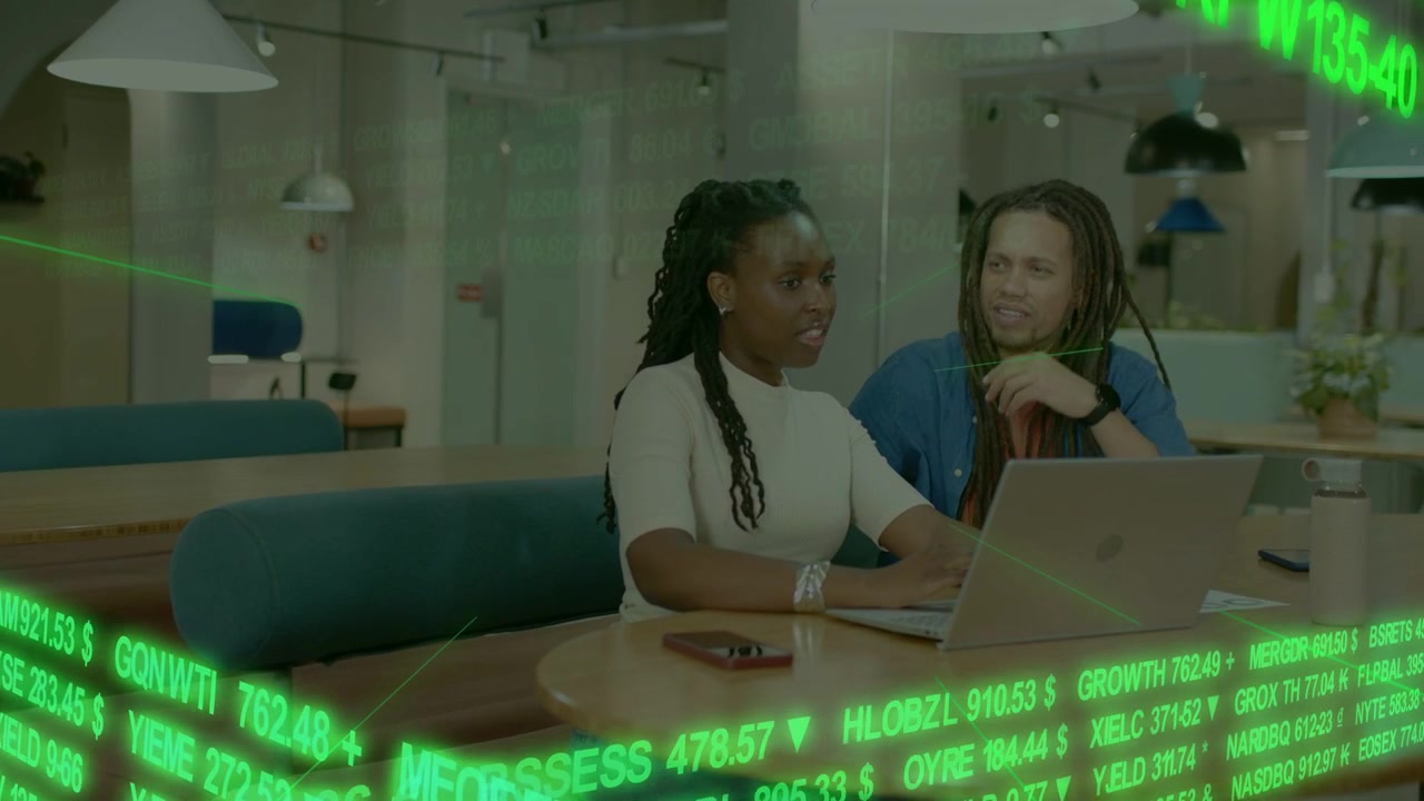 Woman typing laptop while man leaning in pointing, reviewing finance charts with green ticker