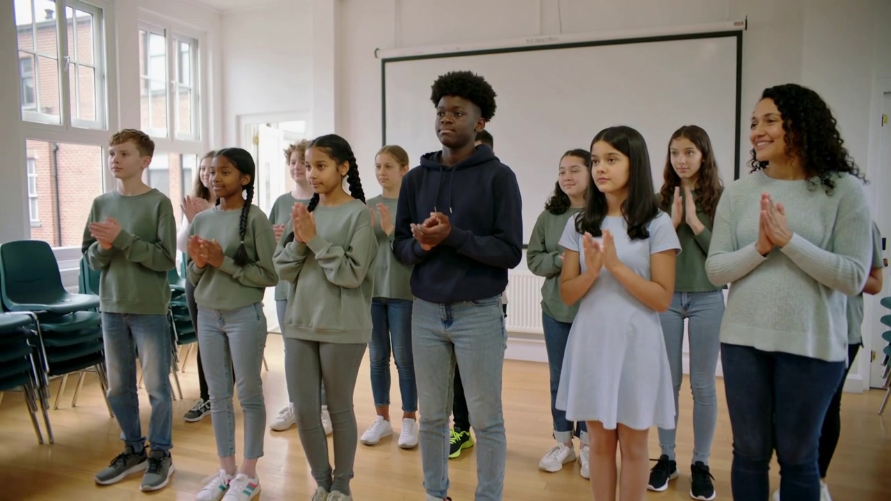 Classe e adulto che applaudono, adolescente centrale in felpa con cappuccio che unisce le mani, praticando il ritmo a scuola, sedie.
