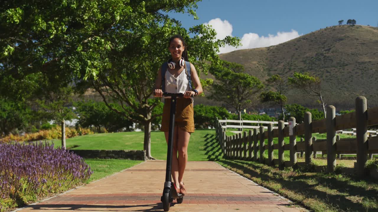 mujer afroamericana con auriculares y mochila montando un scooter en el parque