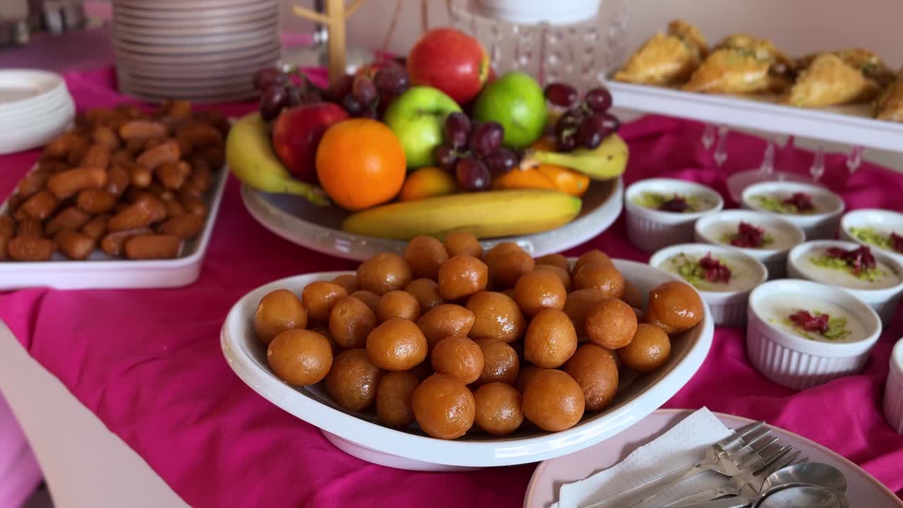 Primo piano di un buffet di dolci tradizionali con gulab jamun, frutta fresca assortita, pasticcini sciroppati e coppette di budino cremoso disposti su un vivace tavolo da catering interno.