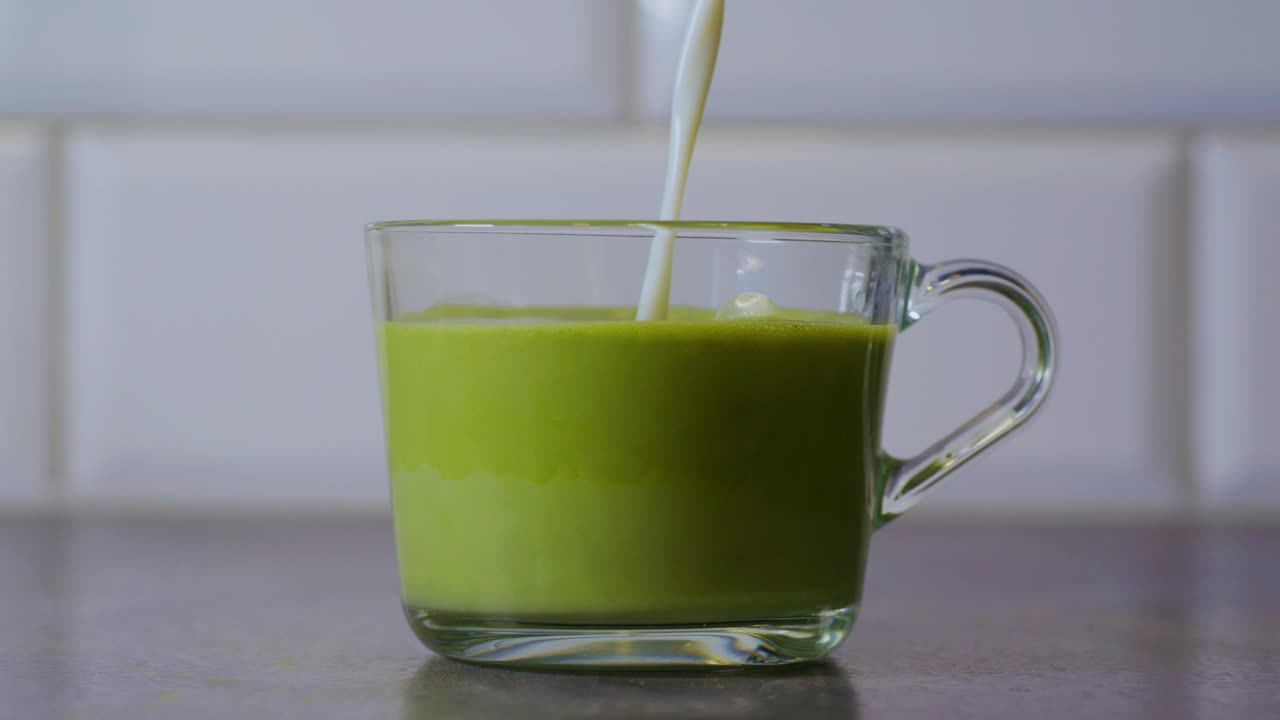 A stream of milk is being poured into a glass cup filled with freshly whisked matcha. The mixture begins to blend, creating a smooth and creamy matcha latte with soft foam on top