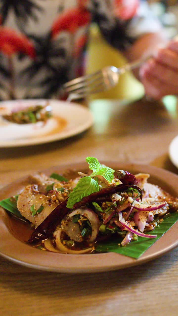 Diner uses fork to eat spicy pork salad, vibrant herbs, natural light, shallow depth