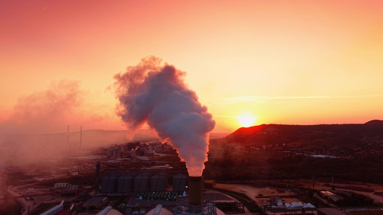 Ciminiera industriale che rilascia una colonna di fumo al tramonto. Il sole che tramonta dietro le colline illumina il fumo denso che sale da una ciminiera di fabbrica in un cielo arancione.