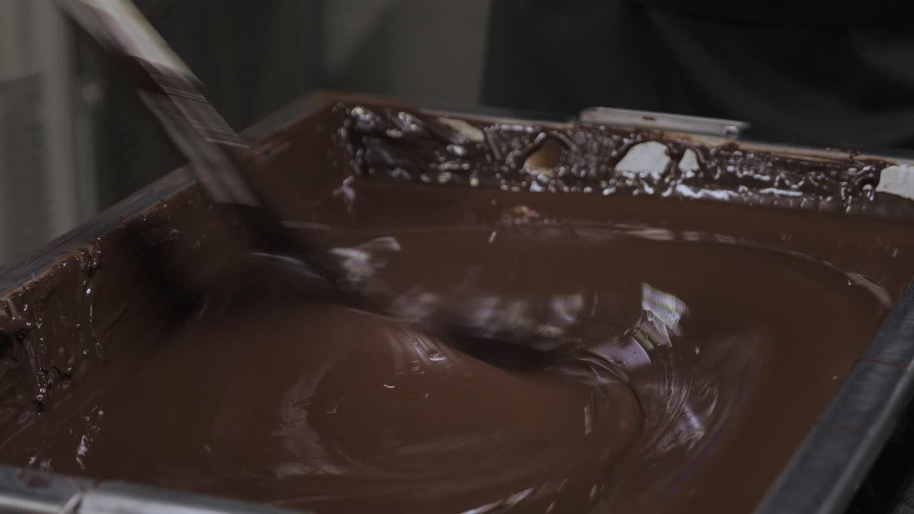 Primo piano di un grande vassoio industriale pieno di cioccolato fondente fuso e lucido, con turbinii
