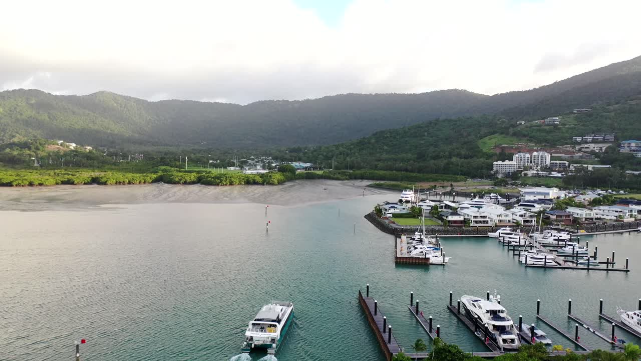 Aerial drone shot in Airlie Beach, Queensland as commercial boat pulls into marina, yachts and houses surrounded by mountain range backdrop.